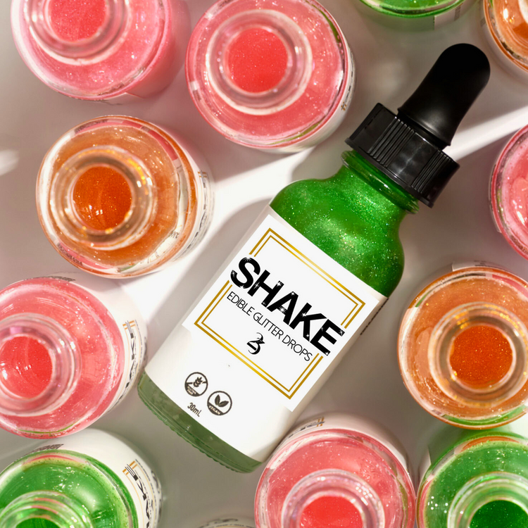 A bottle of SHAKE Edible Glitter Drops surrounded by various colored liquid containers, with the brand name displayed prominently on the bottle.