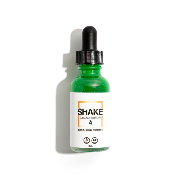 This image displays the SHAKE Edible Glitter Drops bottle, surrounded by various beverage containers, suggesting the product's use. The central focus is on a green bottle with a clean, modern design labeled "SHAKE Edible Glitter Drops," indicating its purpose for enhancing beverages with a touch of sparkle.