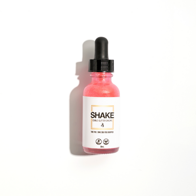 This image displays the SHAKE Edible Glitter Drops bottle, surrounded by various beverage containers, suggesting the product's use. The central focus is on a green bottle with a clean, modern design labeled "SHAKE Edible Glitter Drops," indicating its purpose for enhancing beverages with a touch of sparkle.