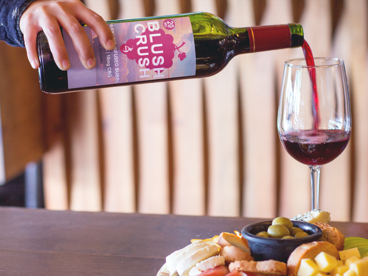 Person pouring red wine from a 'Blush Crush' bottle into a glass with a snack platter on a wooden table.