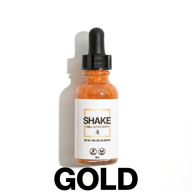 A bottle of SHAKE Edible Glitter Drops surrounded by various colored liquid containers, with the brand name displayed prominently on the bottle.