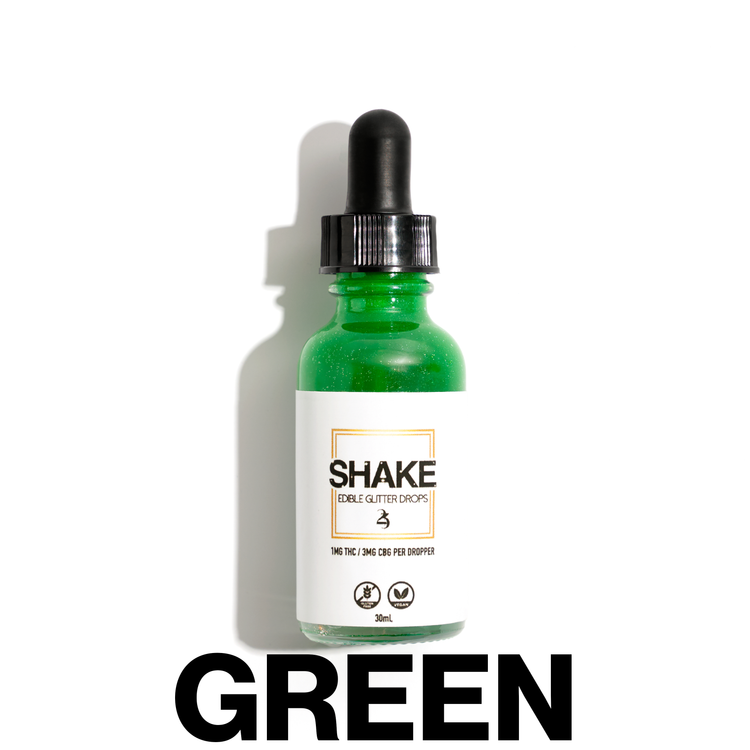 A bottle of SHAKE Edible Glitter Drops surrounded by various colored liquid containers, with the brand name displayed prominently on the bottle.