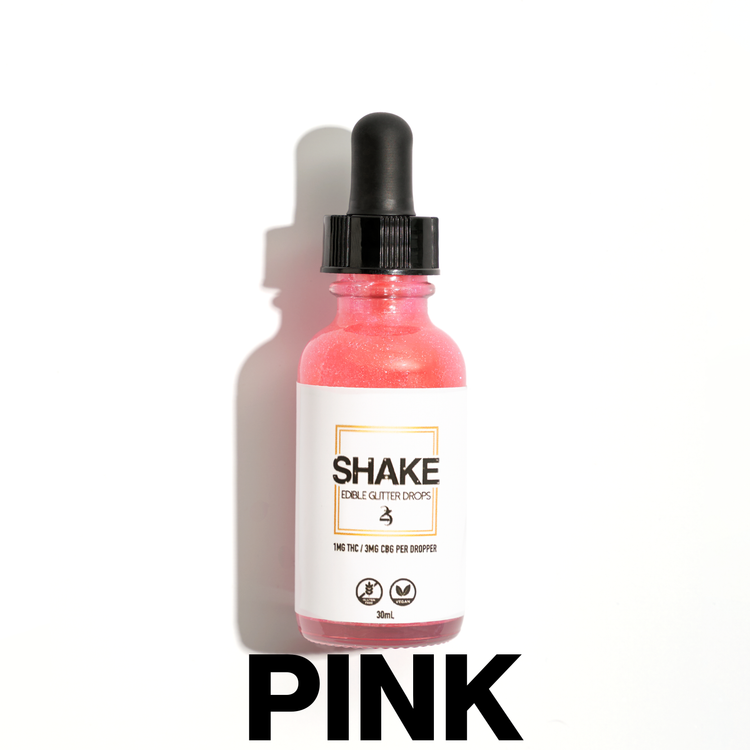 A bottle of SHAKE Edible Glitter Drops surrounded by various colored liquid containers, with the brand name displayed prominently on the bottle.