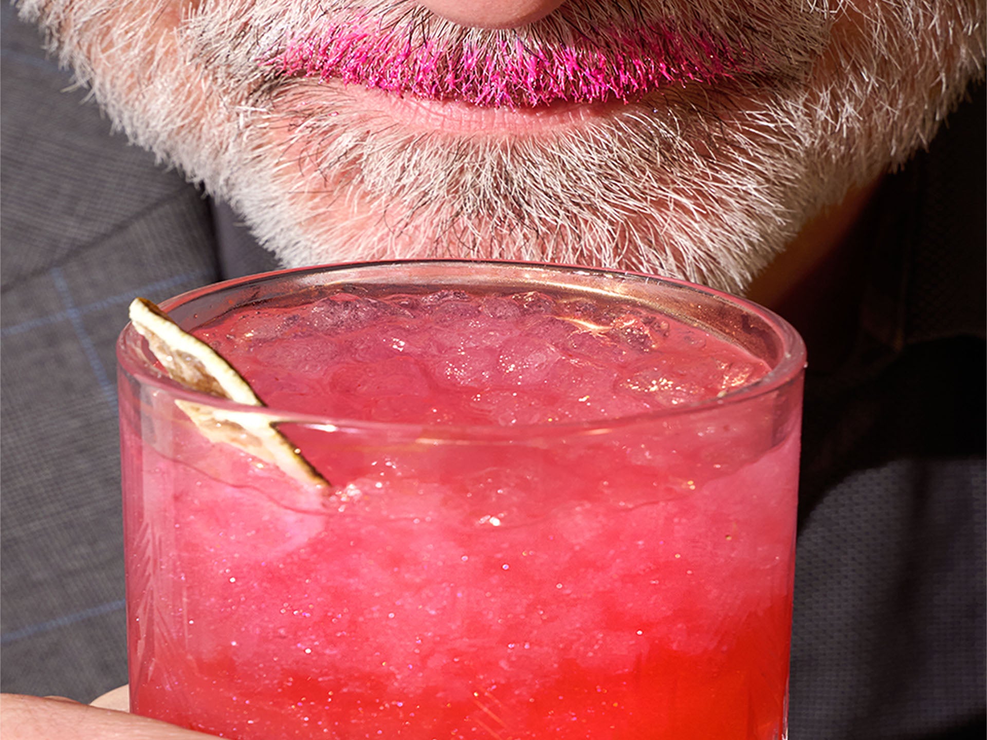 Close-up of a pink cocktail with a lemon twist held by a person with a beard and SHAKE THC infused edible glitter drops.