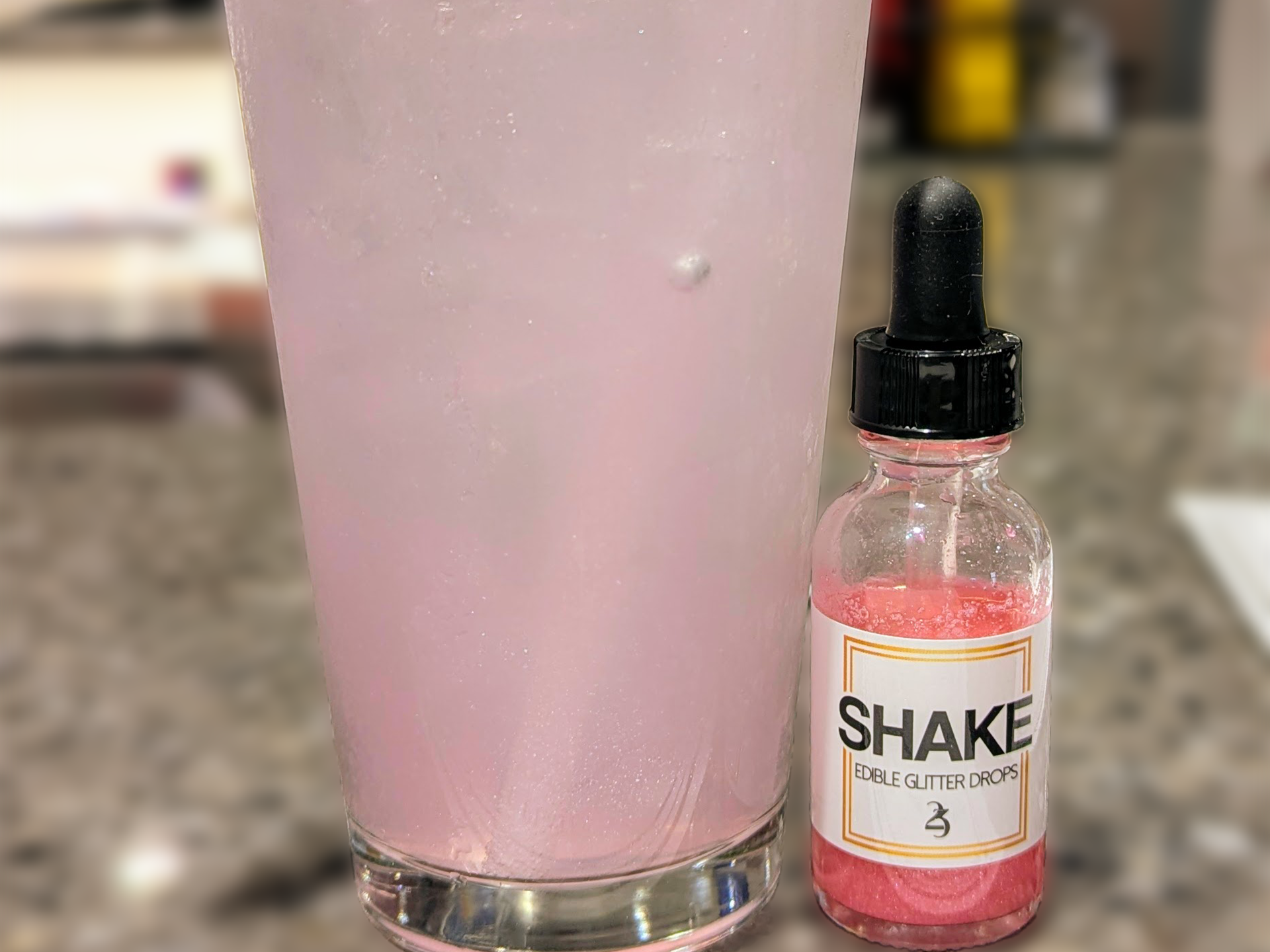 Bottle of 'Shake' edible glitter drops next to a pink drink in a glass.