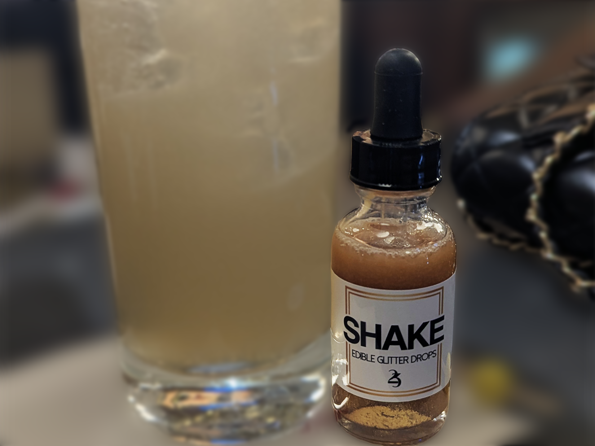 Bottle of 'SHAKE' edible glitter drops with a blurred glass in the background