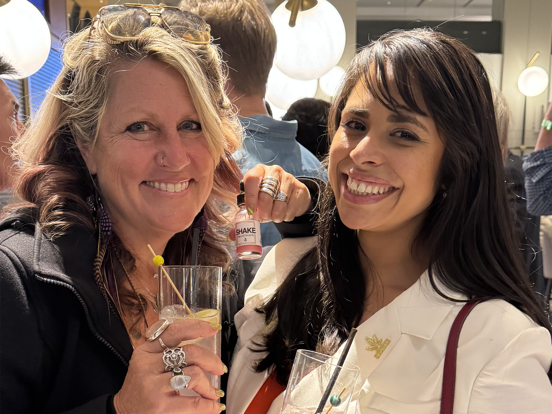 Two women smiling and holding THC drinks infused with SHAKE edible glitter in a social setting.