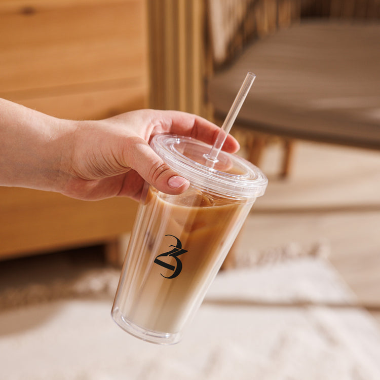 Hand holding a clear cup with a lid and straw, containing a creamy beverage, against a blurred indoor background.