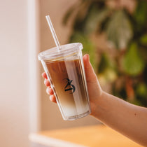 Hand holding a clear cup with iced coffee and a straw, blurred greenery in the background